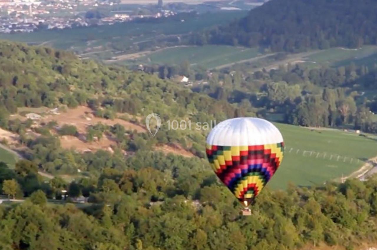 Montgolfière colorée survolant un paysage verdoyant avec collines, champs et quelques habitations au loin.