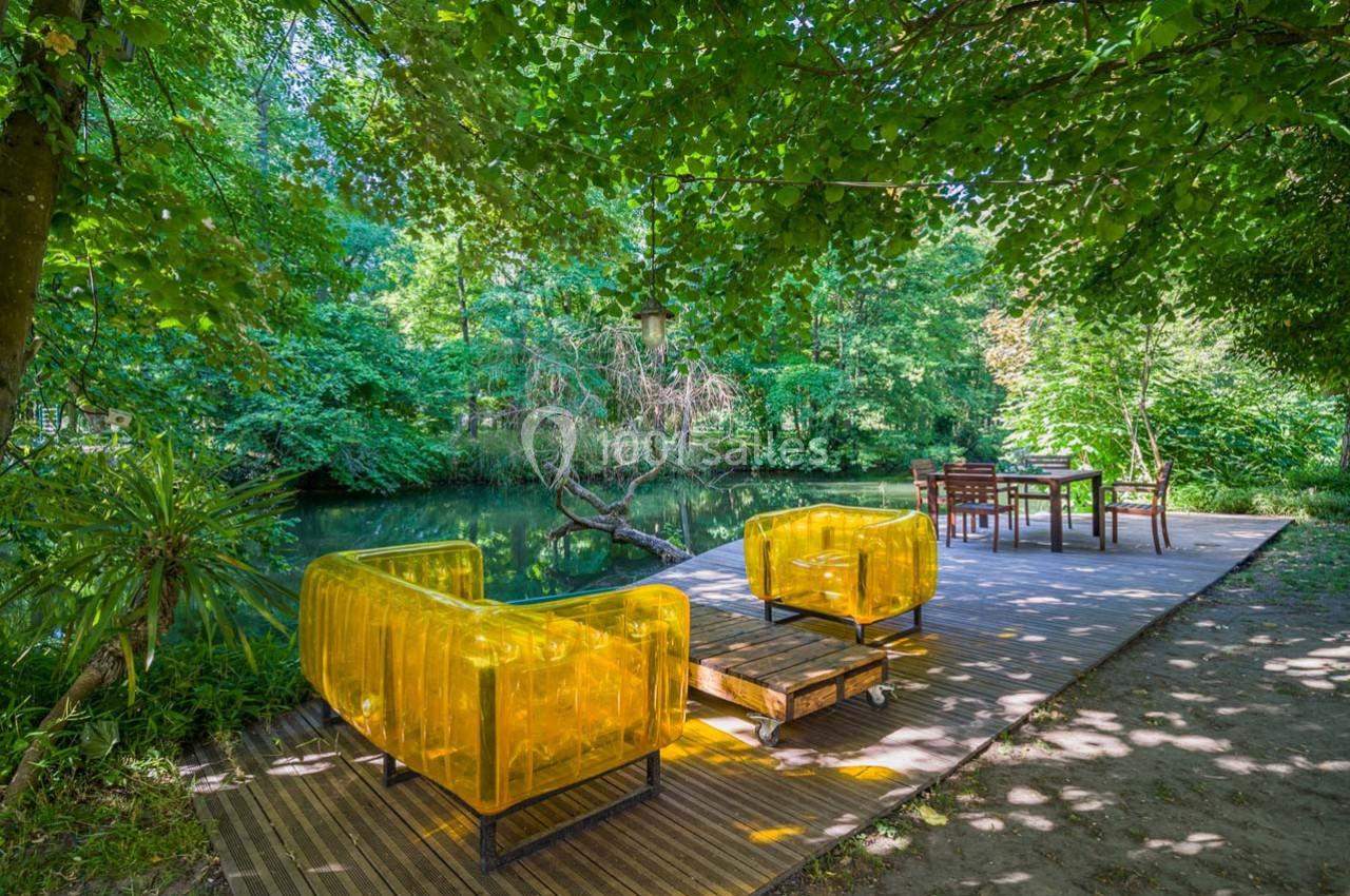 Terrasse en bois au bord d’un étang, avec fauteuils transparents jaunes et table entourée de chaises sous des arbres.