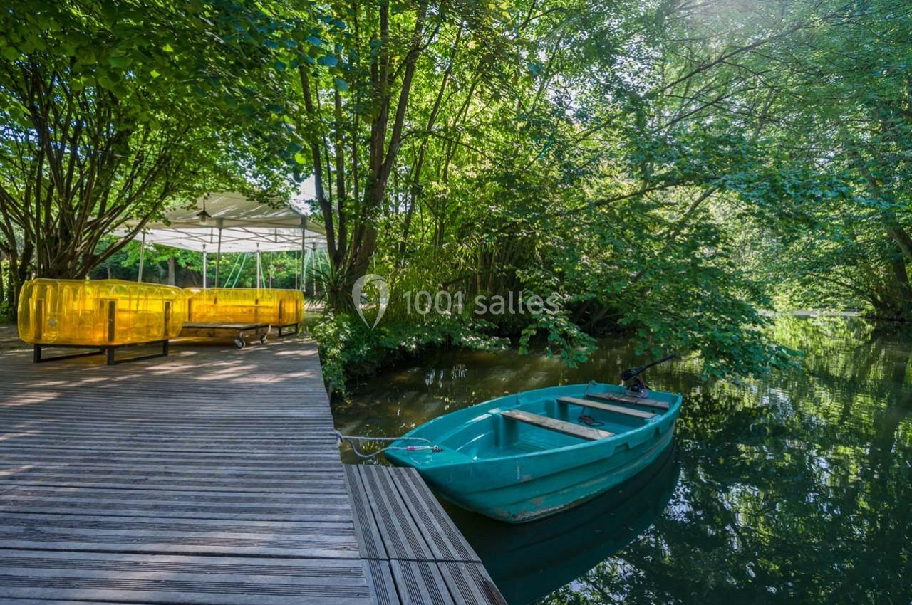 Barque bleue amarrée près d'un ponton en bois, entourée de végétation luxuriante et d'un coin détente avec des sièges jaunes.