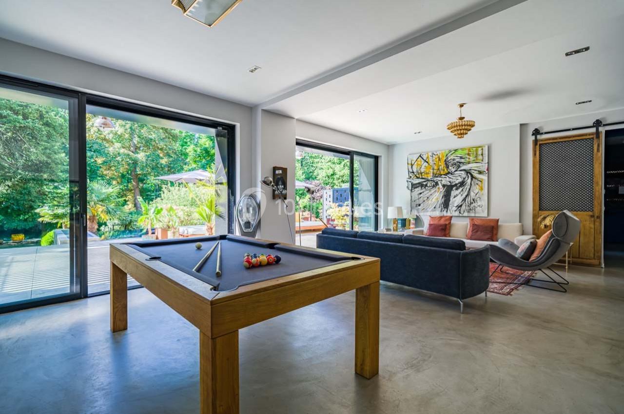 Salle de séjour lumineuse avec table de billard, canapé, œuvres d'art et grandes baies vitrées donnant sur un jardin.