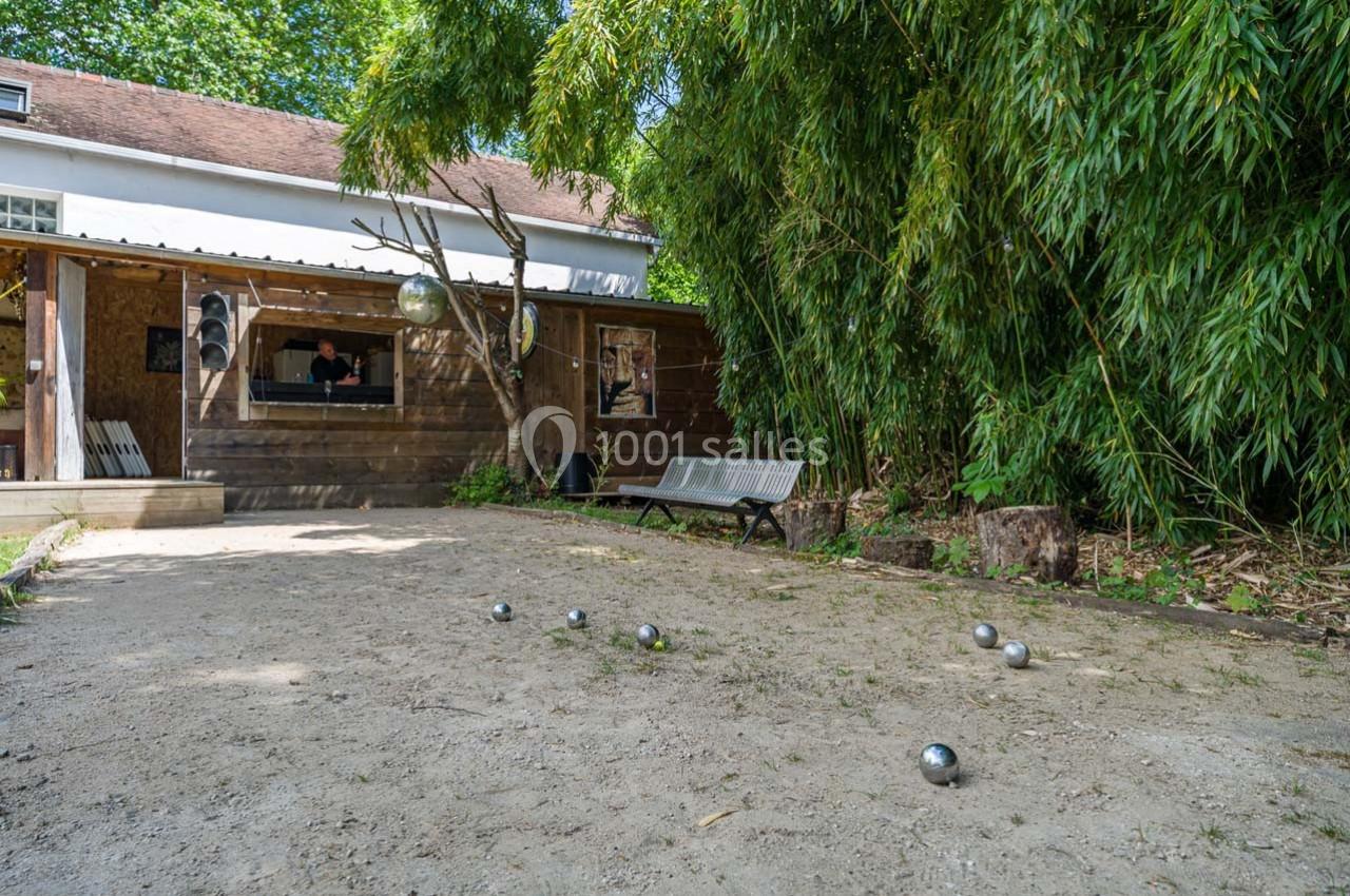 Terrain de pétanque en plein air avec des boules éparpillées, bordé de bambous et d'un bâtiment en bois.