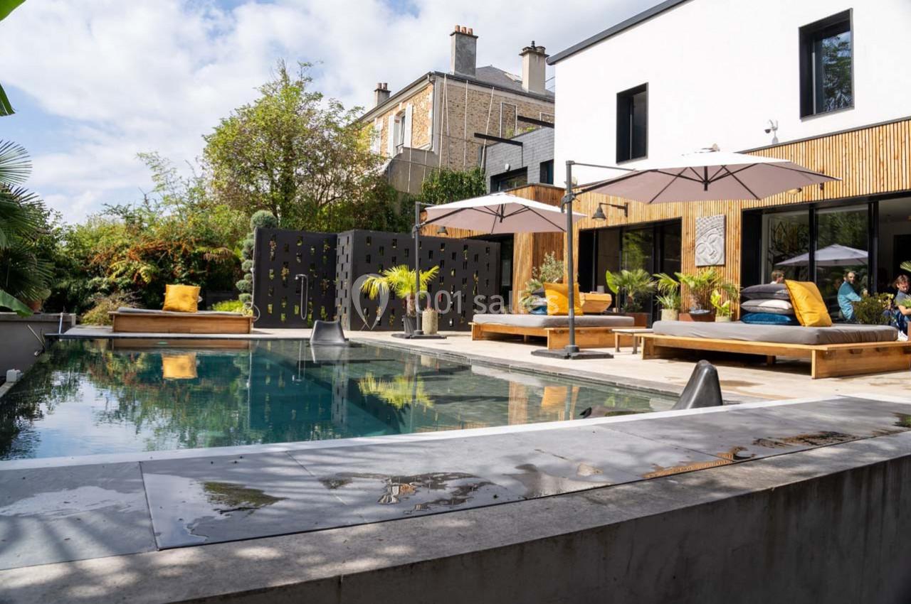 Piscine moderne entourée de terrasses en bois, mobilier extérieur et végétation, devant une maison contemporaine.