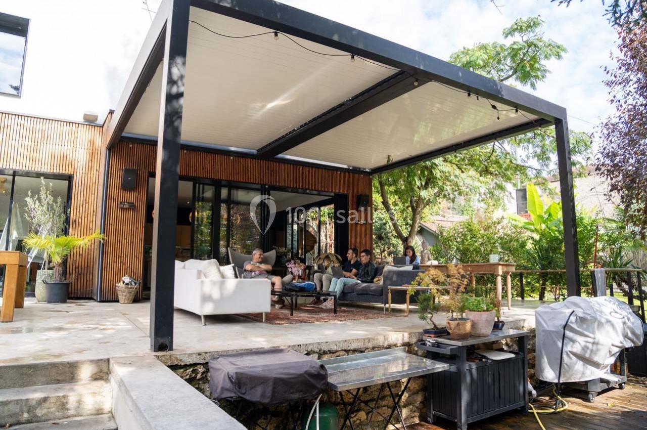 Terrasse moderne avec pergola bioclimatique, salon extérieur et groupe de personnes assises dans un jardin verdoyant.