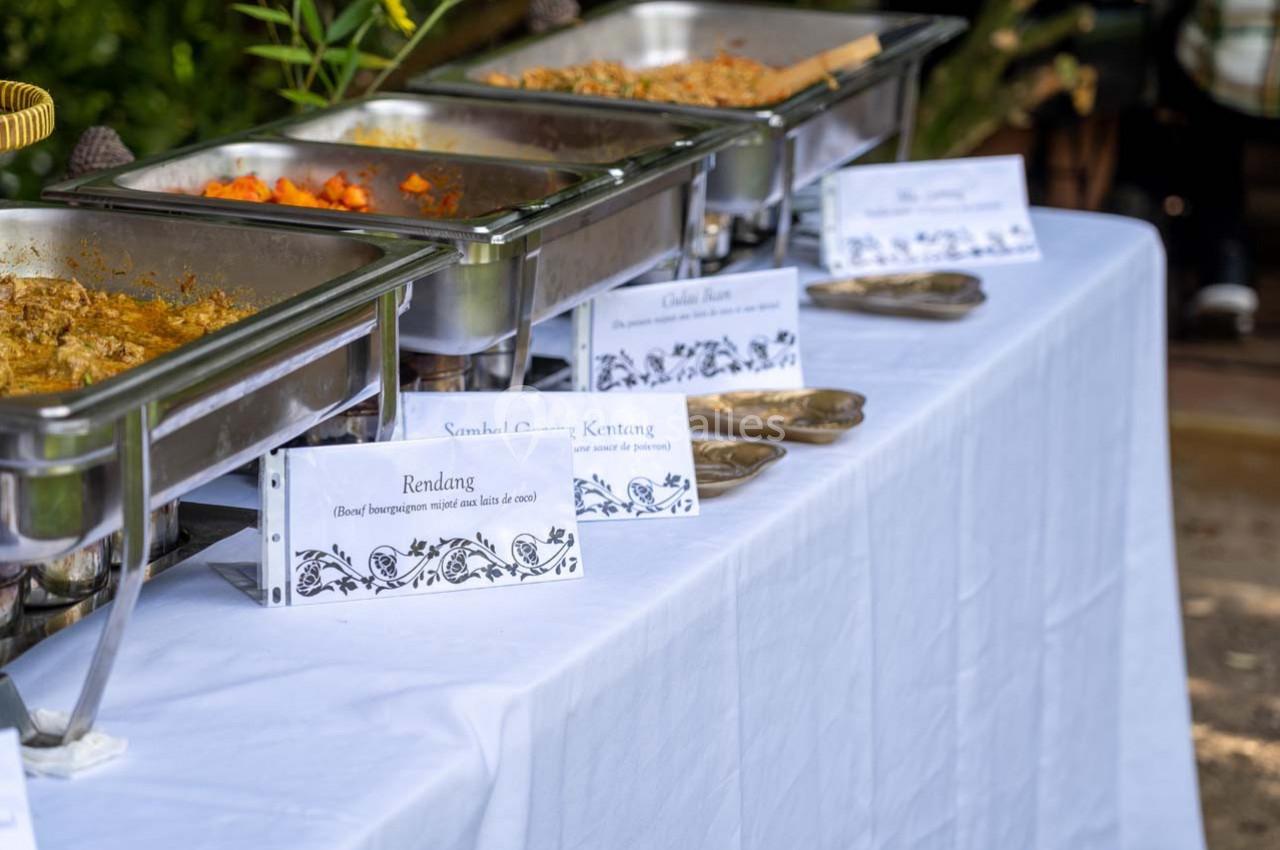 Buffet avec plats chauds présentés dans des bacs en inox, accompagnés d'étiquettes descriptives sur une nappe blanche.