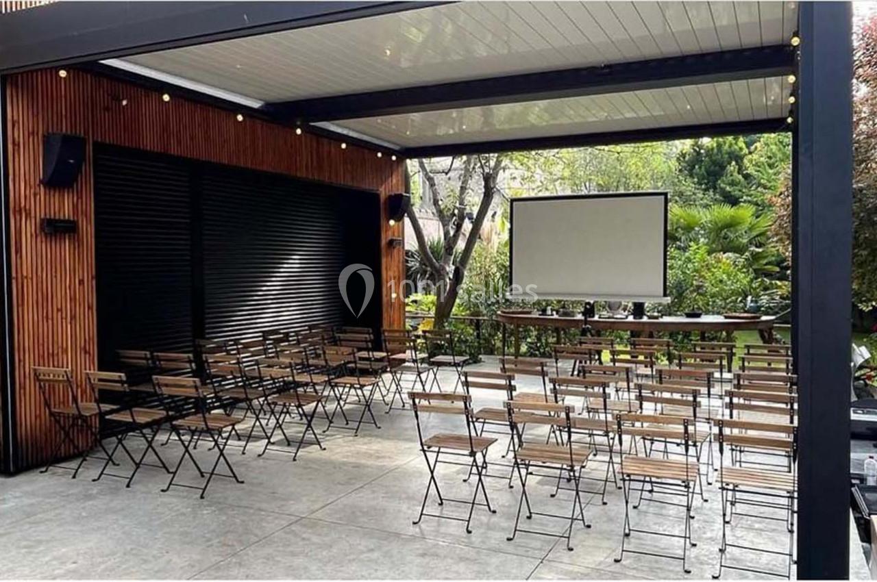 Salle extérieure aménagée avec des chaises pliantes, un écran de projection et un cadre verdoyant en arrière-plan.