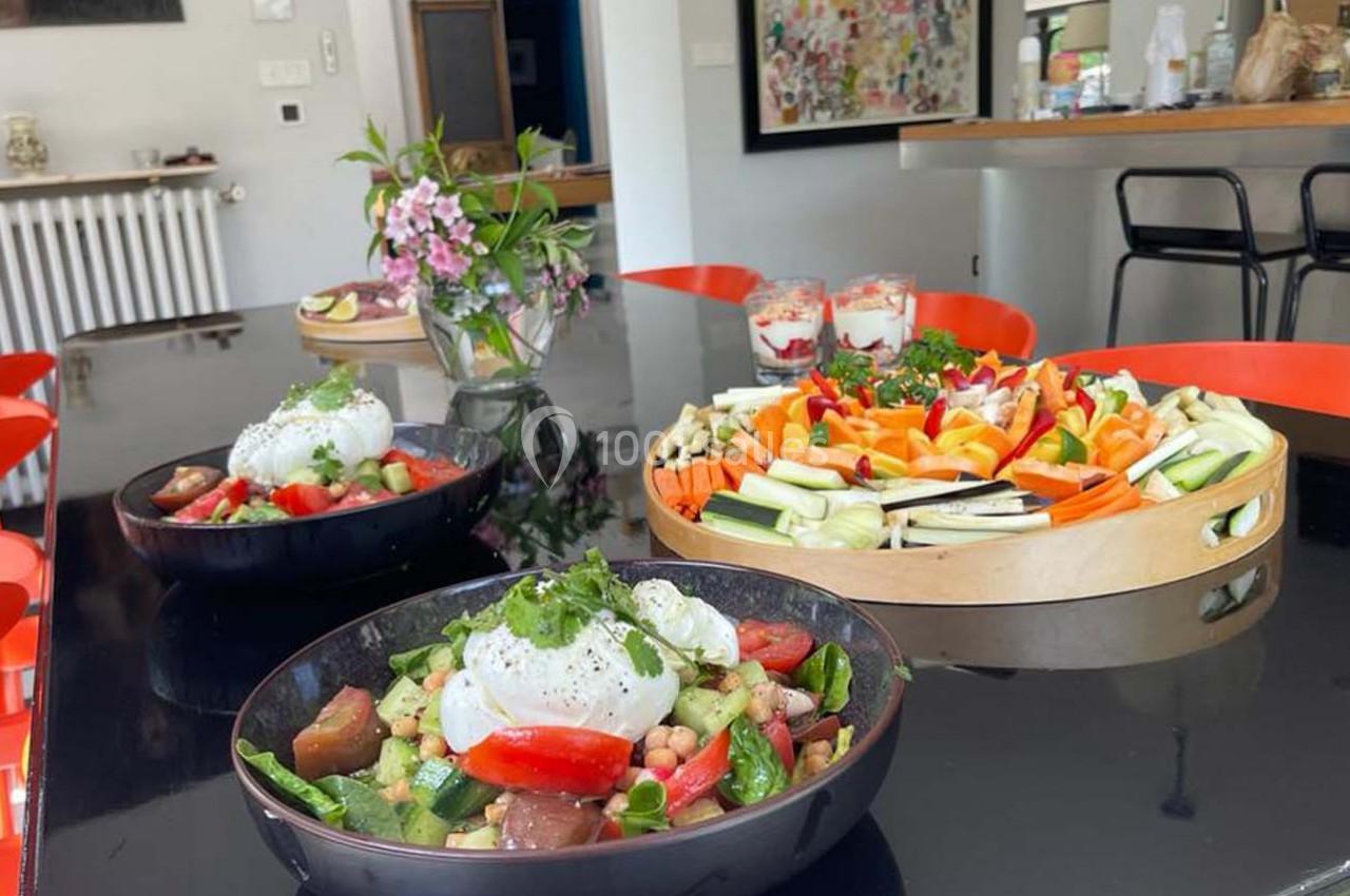 Assiette de salade colorée avec burrata au premier plan, plateau de crudités et fleurs en arrière-plan sur une table noire.