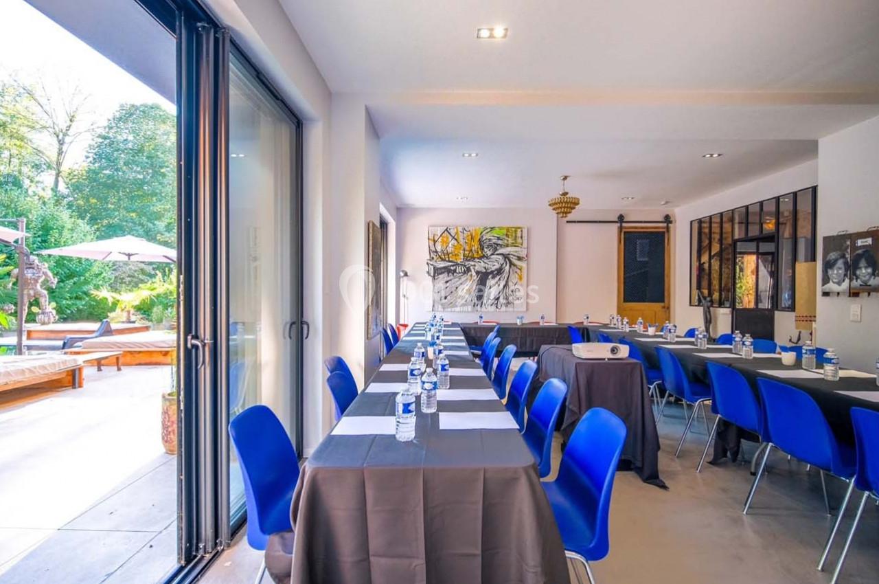 Salle de réunion lumineuse avec tables alignées, chaises bleues, vue sur une terrasse et jardin arboré.