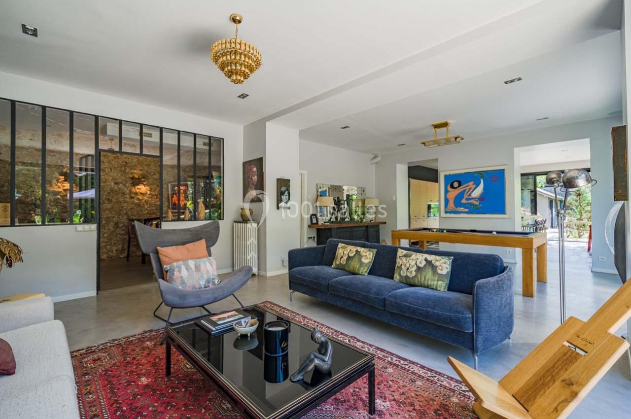 Salon lumineux avec canapé bleu, fauteuil, table basse, tapis rouge, verrière et vue sur une salle à manger ouverte.