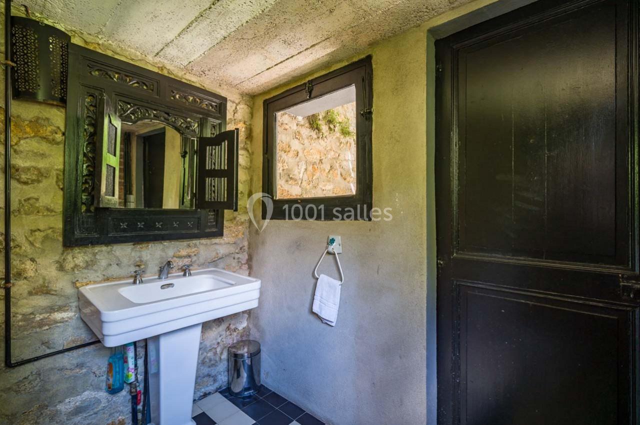 Salle de bain rustique avec lavabo, miroir encadré, mur en pierre, fenêtre et porte en bois sombre.