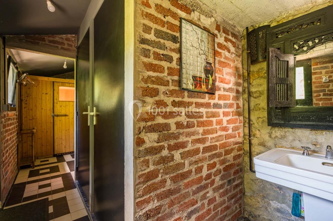 Mur en briques apparentes avec un lavabo blanc, un miroir encadré en bois et une porte en bois au fond.
