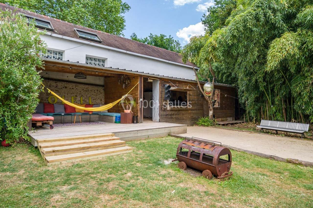 Espace extérieur avec terrasse en bois, hamac, banc, barbecue et végétation dense autour d'une maison à étage.