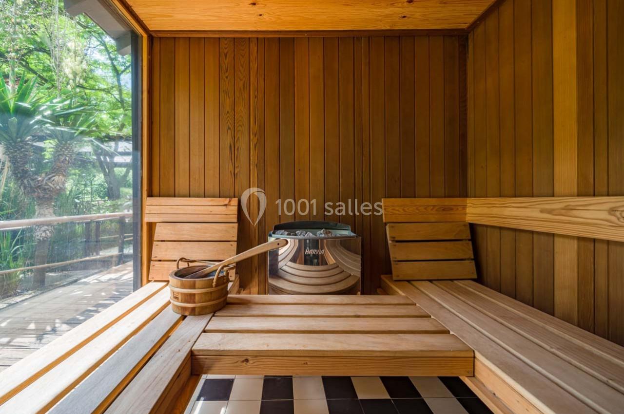 Intérieur d'un sauna en bois avec bancs, seau en bois et poêle, vue sur un jardin à travers une grande fenêtre.