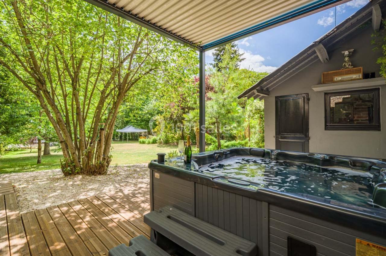 Jacuzzi extérieur sur une terrasse en bois, entouré de verdure avec un jardin arboré en arrière-plan.