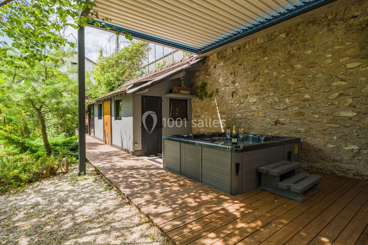 Terrasse en bois avec un jacuzzi, adossée à un mur en pierre, entourée de verdure et partiellement couverte.
