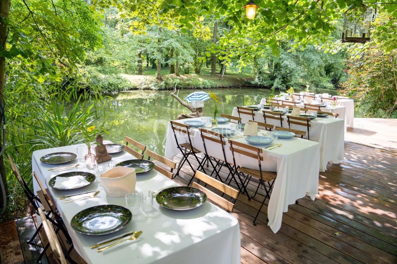 Tables dressées avec nappes blanches et vaisselle, installées sur une terrasse en bois au bord d'un étang entouré de verdure.