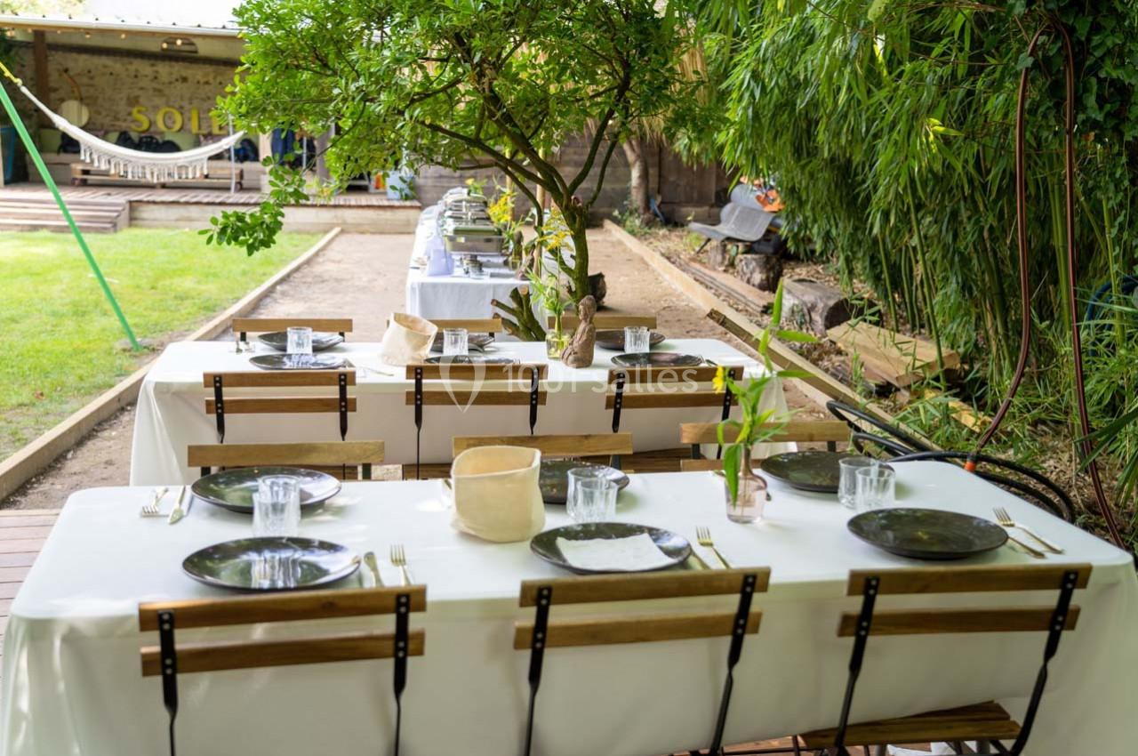 Tables en bois dressées avec nappes blanches, assiettes et verres, disposées en extérieur sous des arbres.
