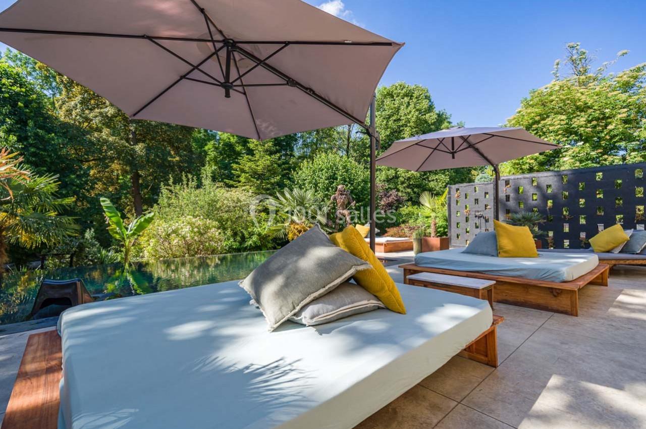 Chaises longues avec coussins sous des parasols, près d'une piscine entourée de végétation luxuriante.
