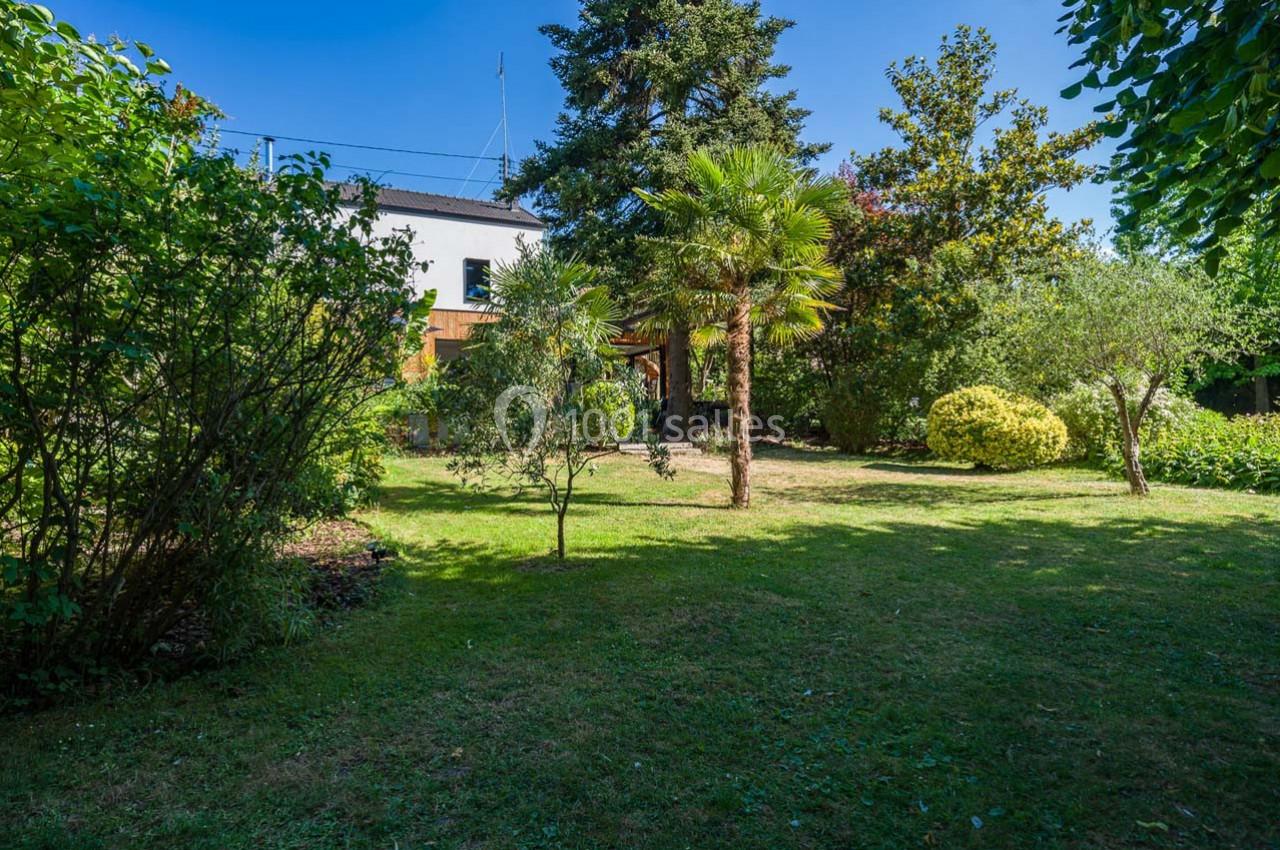 Jardin verdoyant avec pelouse, arbres et arbustes, entouré de végétation, maison blanche visible en arrière-plan.