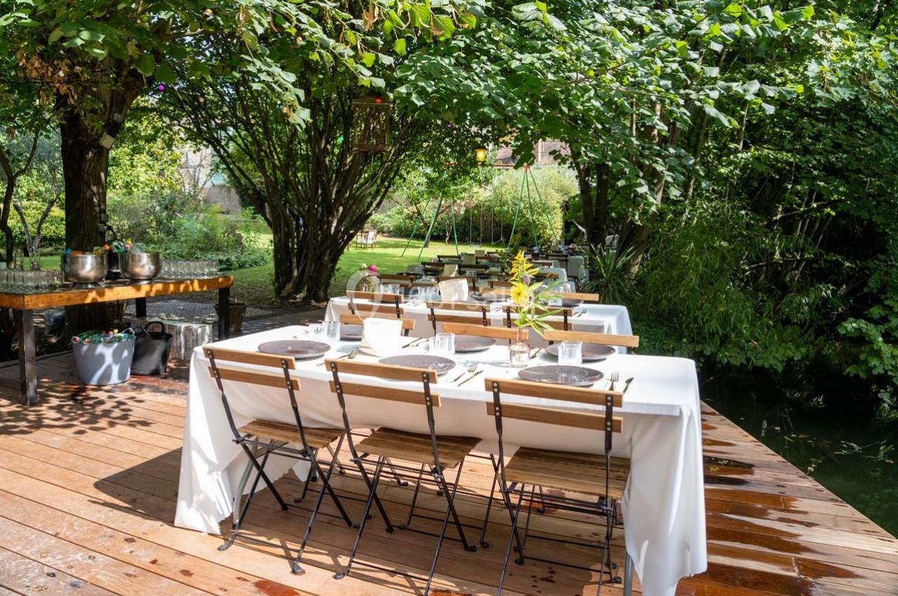 Tables dressées avec nappes blanches sur une terrasse en bois, entourée de verdure et d'arbres dans un jardin.
