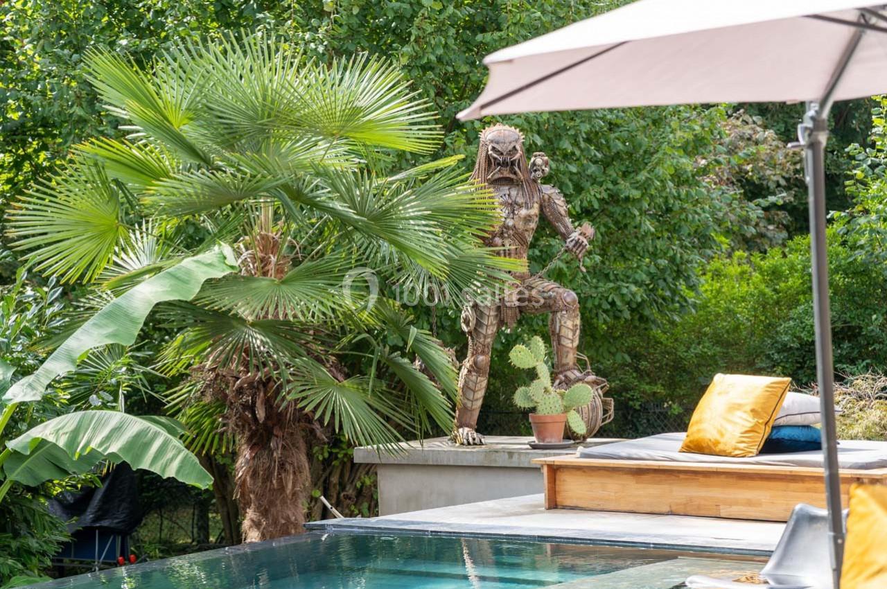 Statue en métal représentant un personnage fantastique, placée près d'une piscine entourée de végétation tropicale.