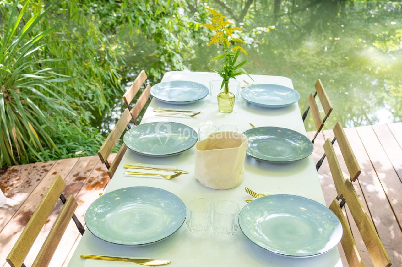 Table dressée en extérieur avec vaisselle bleue, couverts dorés, vase de fleurs jaunes et vue sur un cours d'eau.