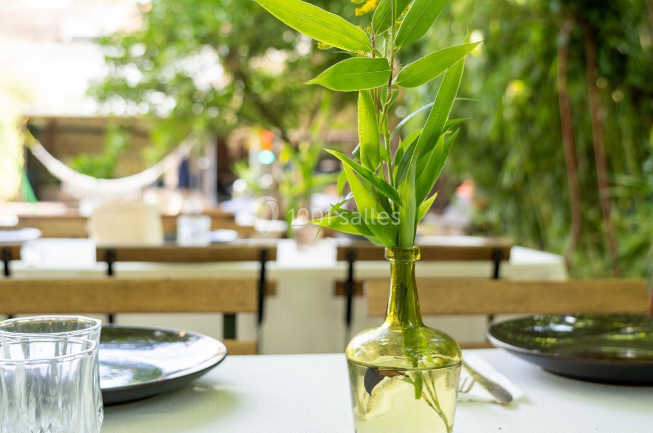 Un vase en verre avec des feuilles vertes posé sur une table dressée en extérieur, entourée de végétation.