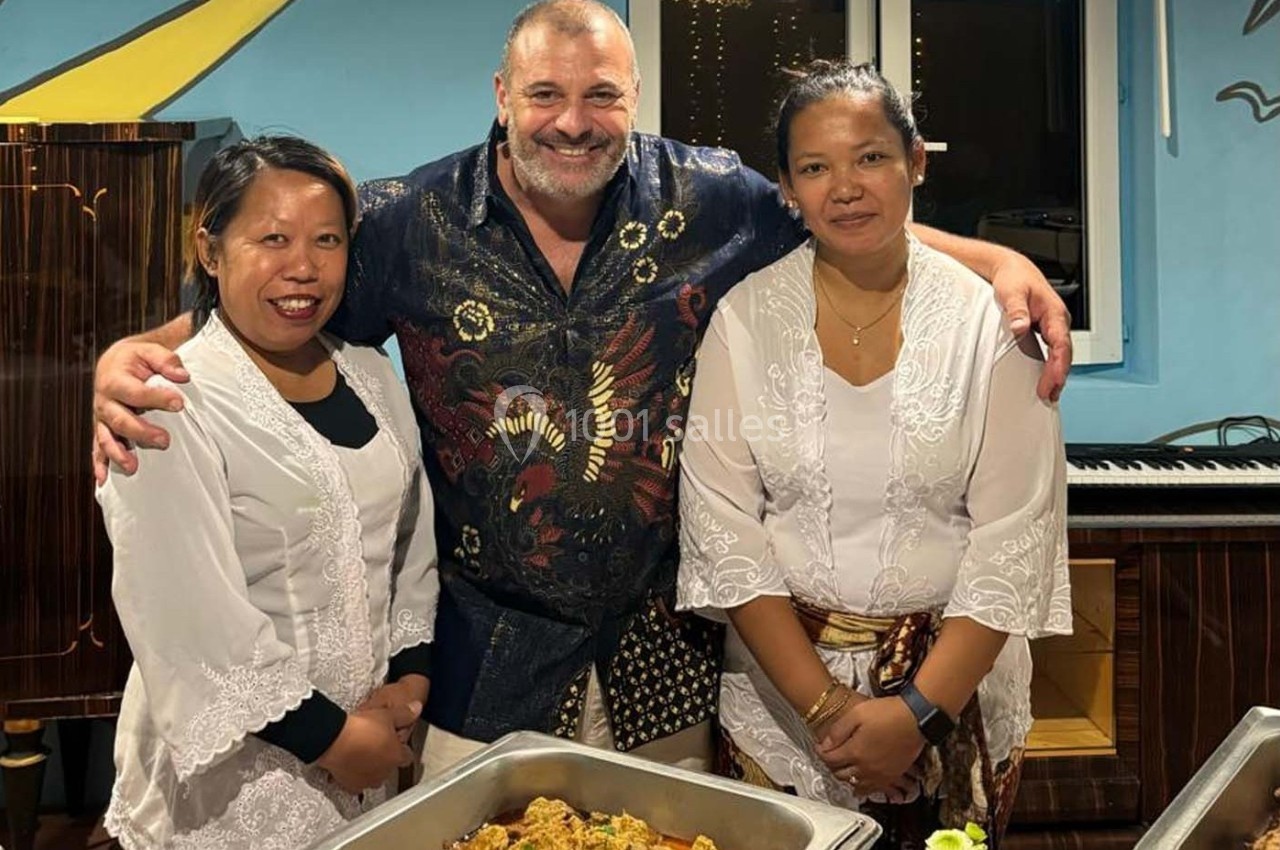 Un homme souriant pose avec deux femmes en tenue traditionnelle devant des plats cuisinés dans un cadre intérieur.