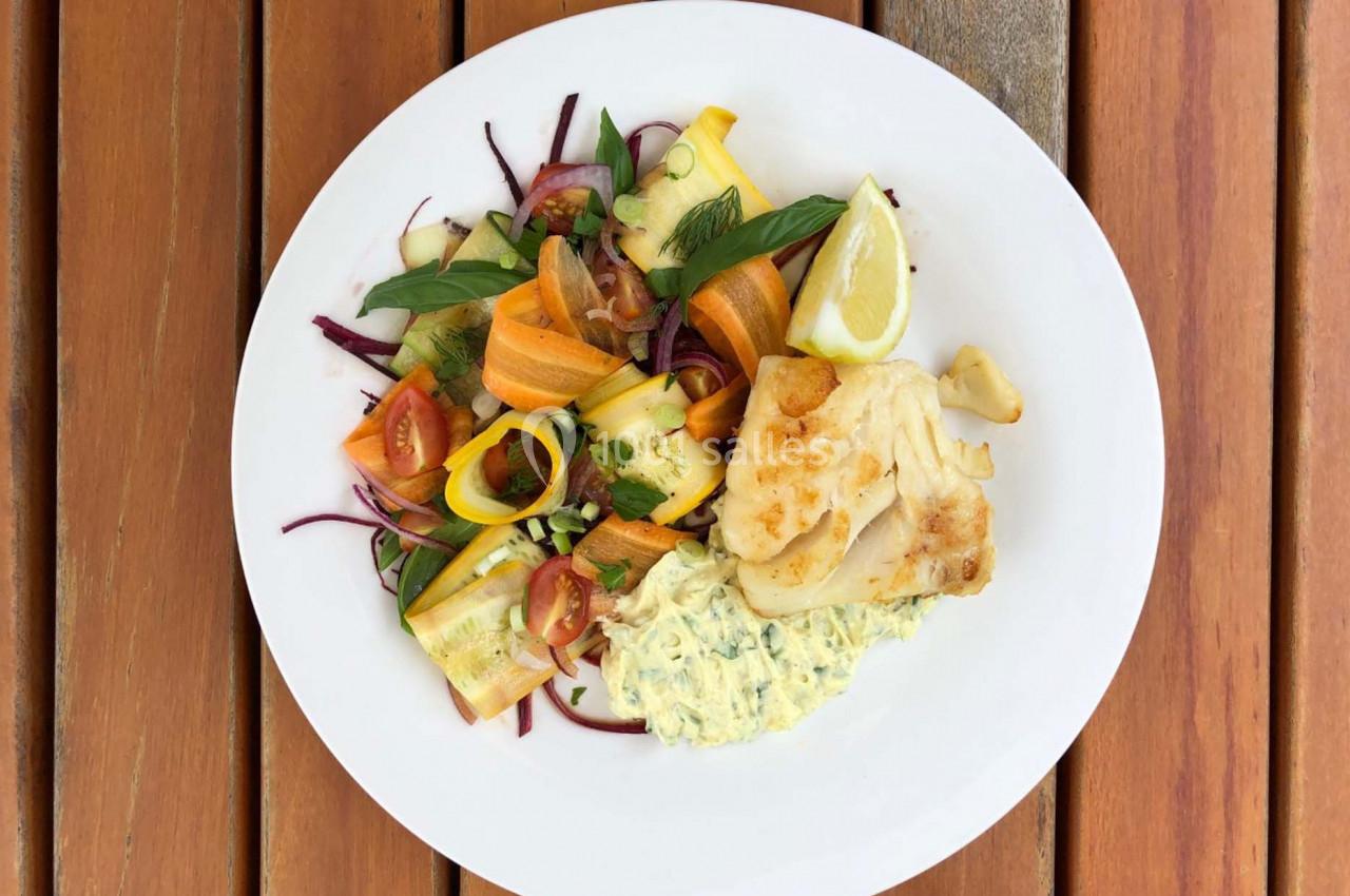 Assiette contenant du poisson grillé, une salade de légumes colorés et une sauce crémeuse, servie sur une table en bois.