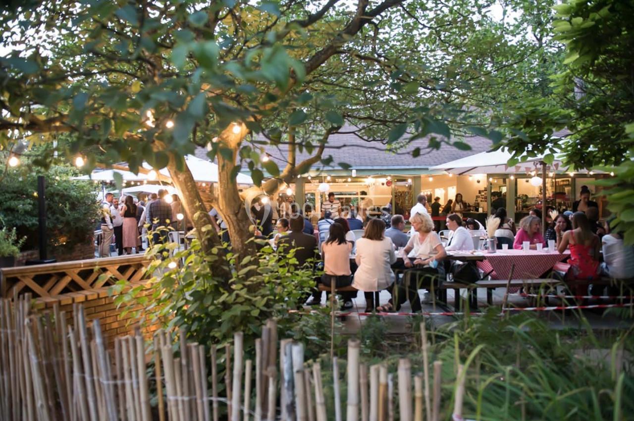 Des personnes dînent en plein air dans un jardin éclairé par des guirlandes lumineuses, entouré de verdure.