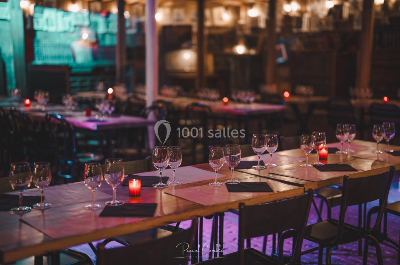 Salle de restaurant avec tables alignées, nappes en tissu, verres à vin et bougies rouges allumées.
