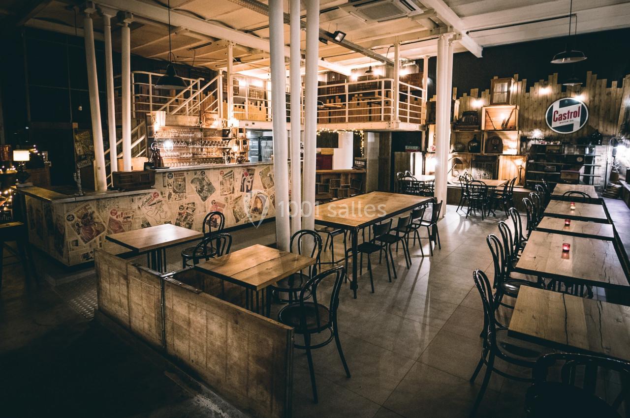Salle de restaurant au style industriel avec tables en bois, chaises noires, bar en arrière-plan et éclairage tamisé.