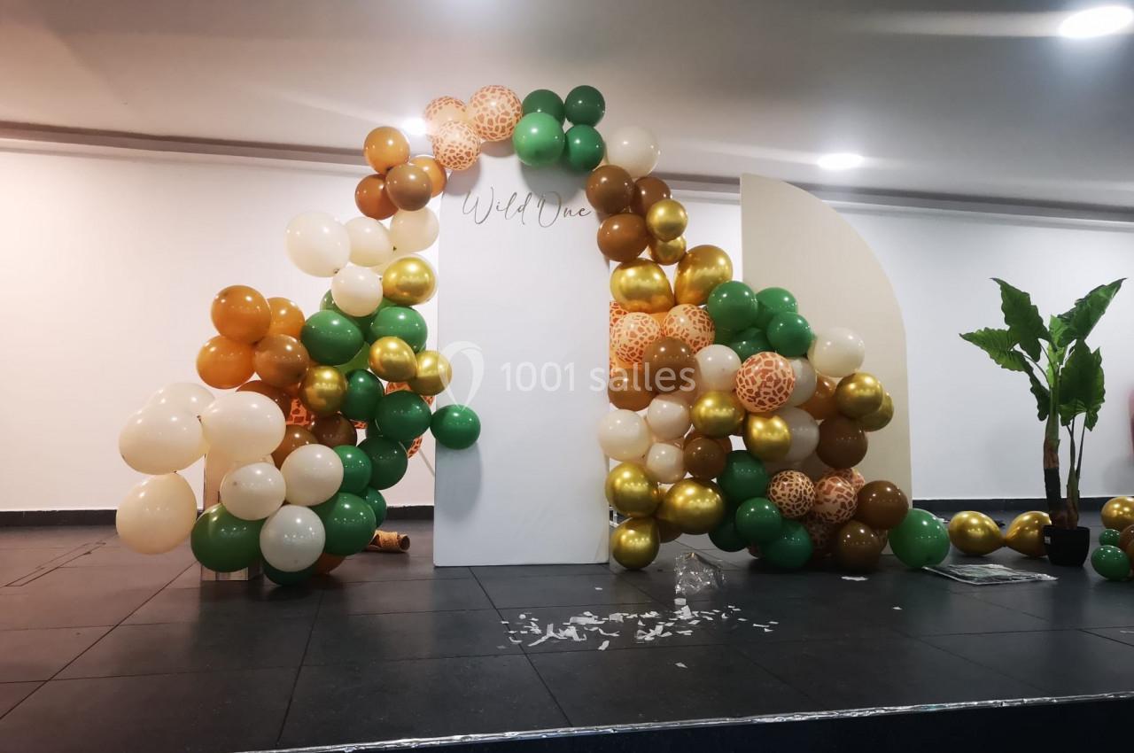 Arche décorative en ballons verts, dorés, blancs et imprimés, installée dans une salle avec un sol sombre.