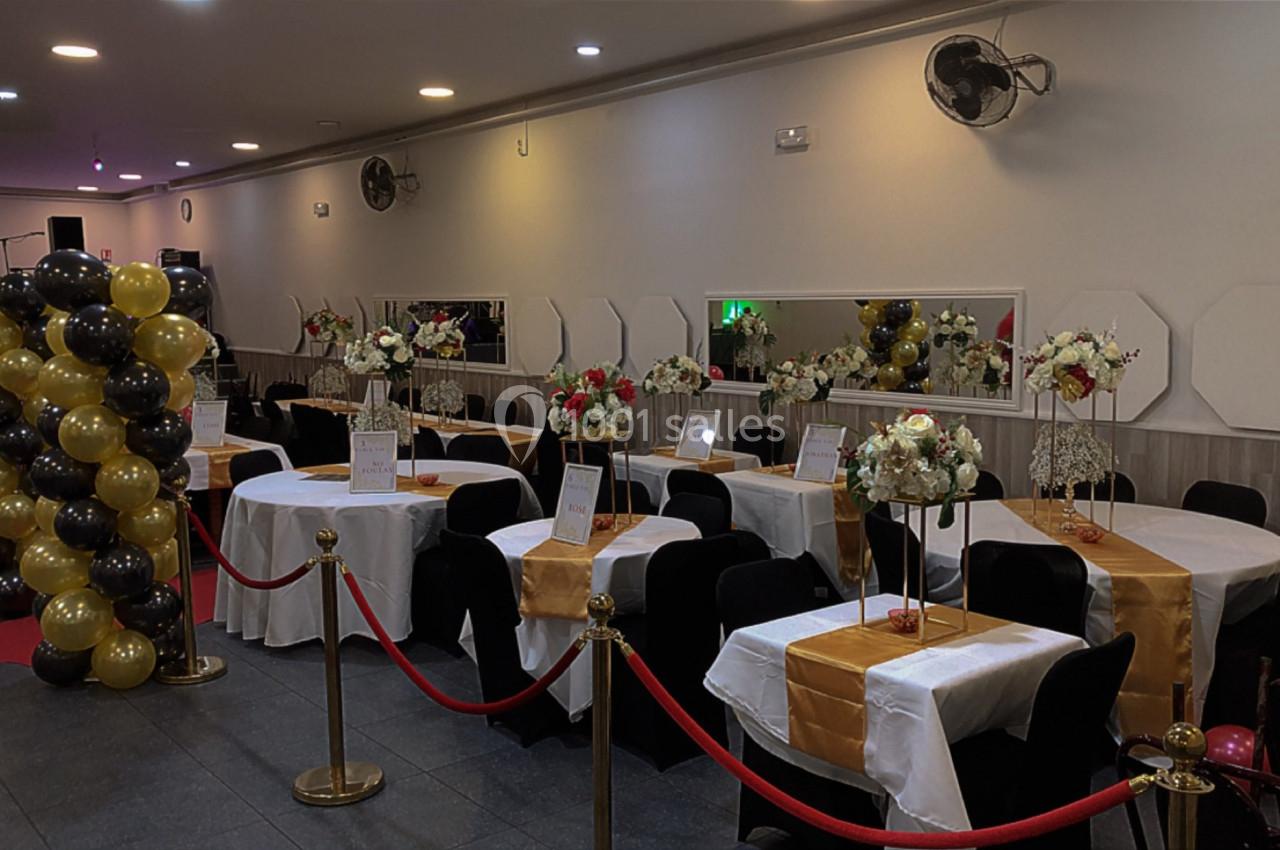 Salle de réception décorée avec des tables dressées, des ballons noirs et dorés, et des fleurs en centre de table.