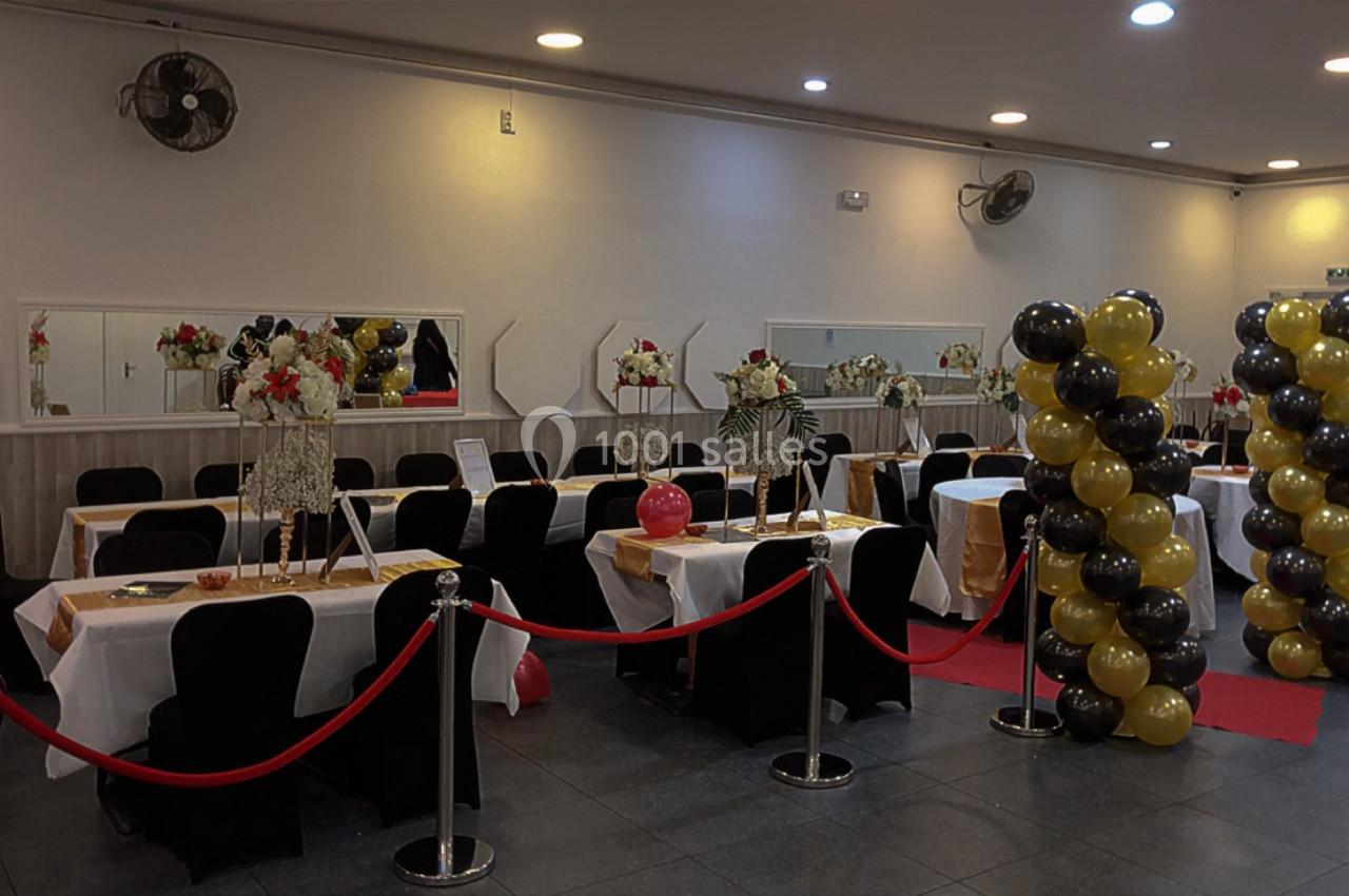 Salle de réception décorée avec des tables dressées, des ballons noirs et dorés, et des cordons rouges.