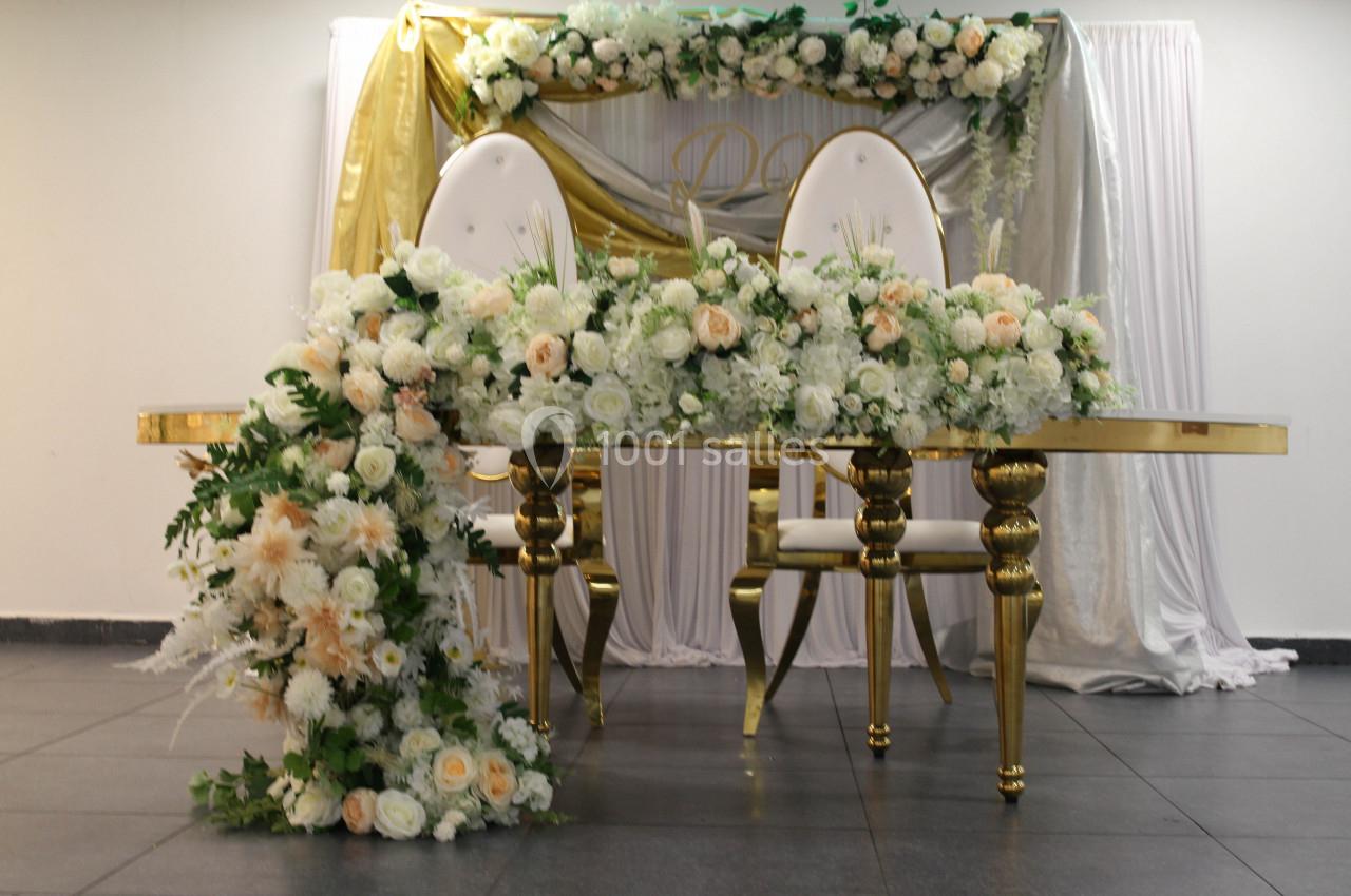 Table décorée pour un mariage avec des fleurs blanches et pêche, chaises dorées et arrière-plan drapé blanc et or.