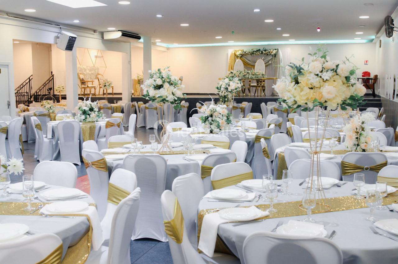 Salle de réception décorée avec des tables rondes, nappes blanches, chemins de table dorés et arrangements floraux.