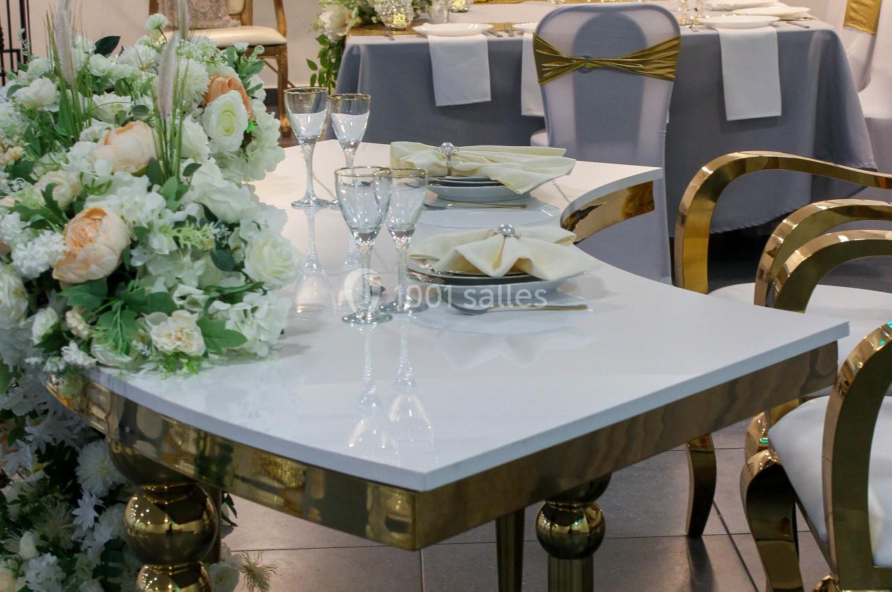 Salle de réception élégante avec tables décorées de fleurs blanches et dorées, vaisselle raffinée et chaises dorées.