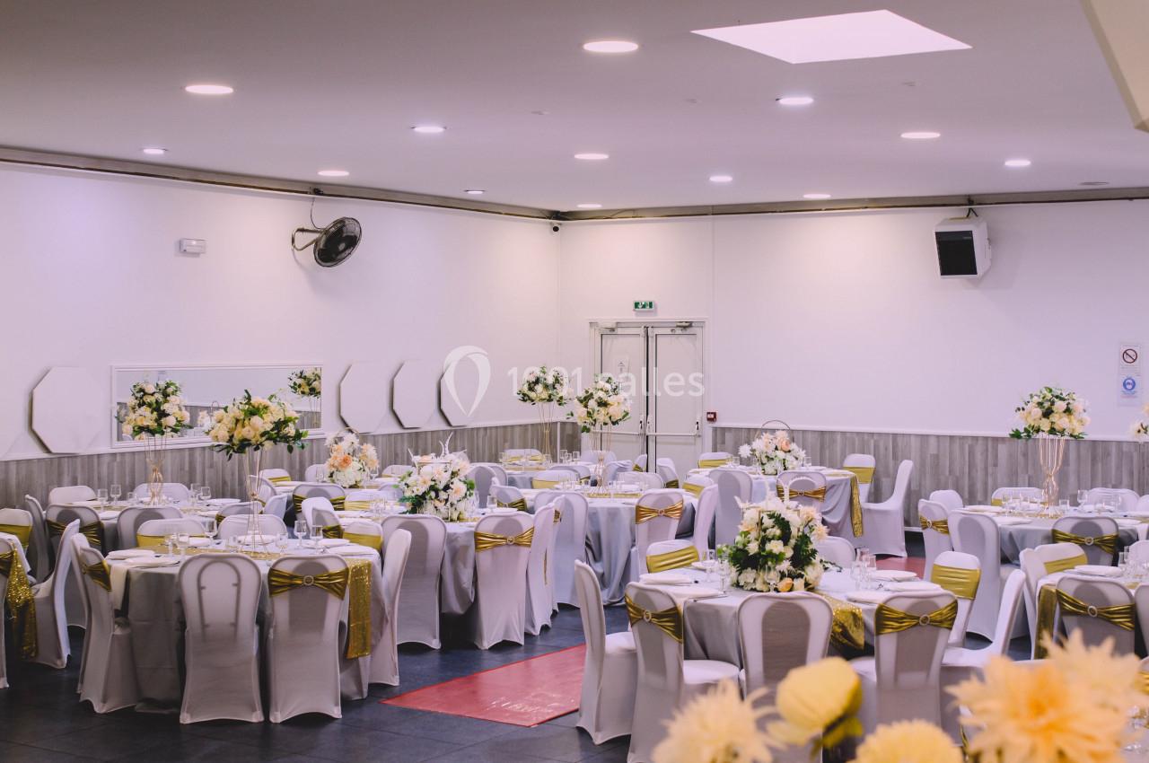 Salle de réception décorée avec des tables rondes, nappes blanches, chaises ornées de rubans dorés et bouquets de fleurs.