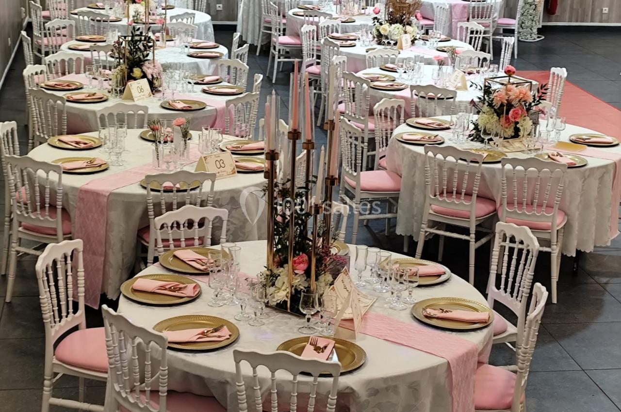 Salle de réception décorée avec des tables rondes ornées de nappes blanches, chemins de table roses et compositions florales.