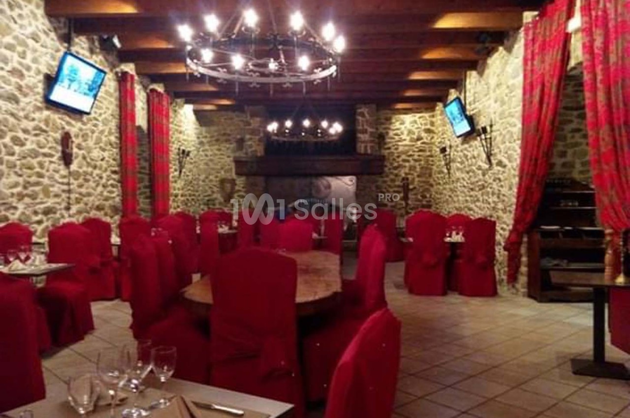 Salle de restaurant avec murs en pierre, chaises rouges, tables dressées et lustres suspendus au plafond en bois.
