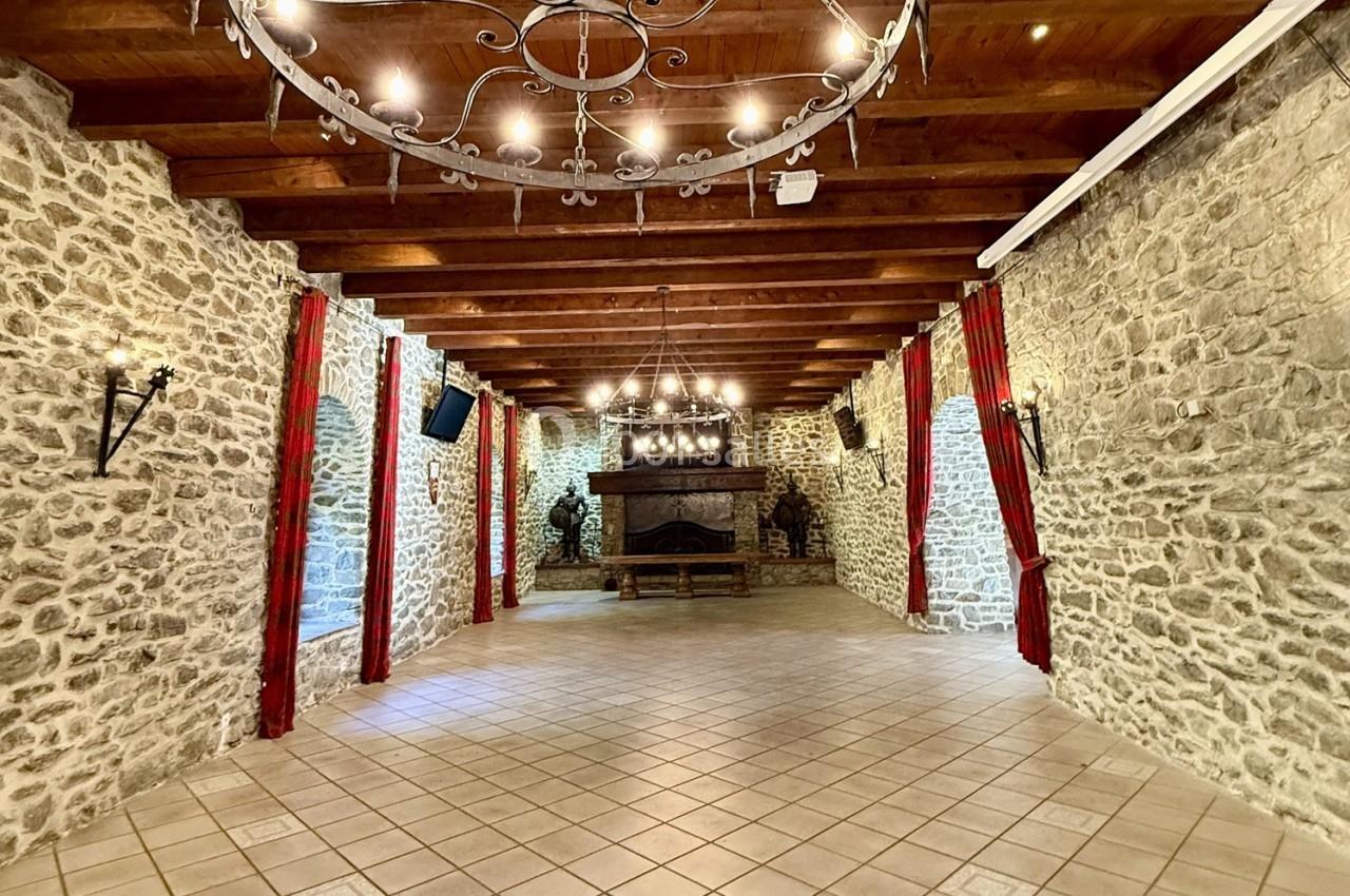 Salle en pierre avec poutres apparentes, cheminée centrale, lustres en fer forgé et rideaux rouges encadrant les murs.