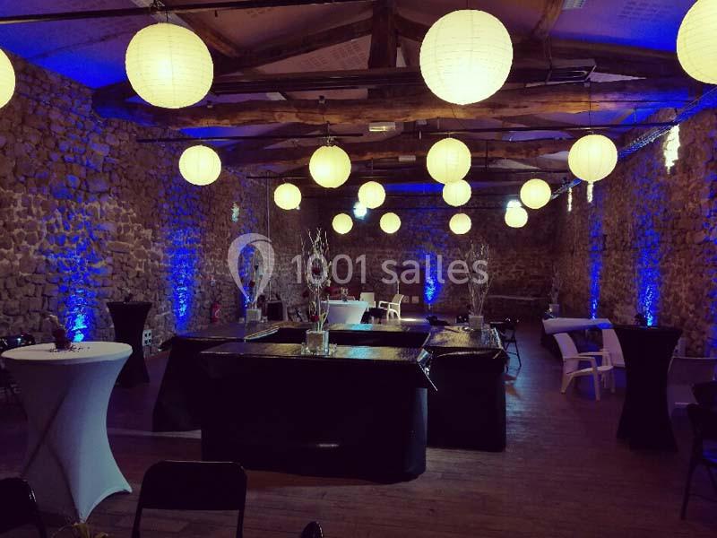 Salle en pierre avec des tables décorées, éclairée par des lanternes suspendues et des lumières bleues.