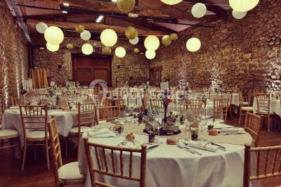 Salle en pierre avec tables rondes dressées pour un événement, décorée de lanternes suspendues et de fleurs.