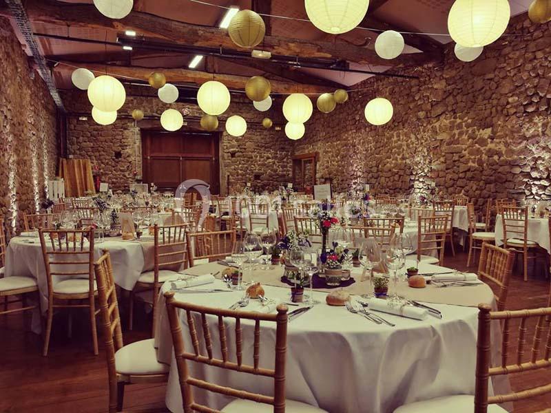 Salle en pierre avec tables rondes dressées pour un événement, décorée de lanternes suspendues et de fleurs.