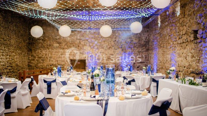 Salle en pierre décorée pour un mariage avec tables rondes dressées, nappes blanches, guirlandes lumineuses et lanternes…