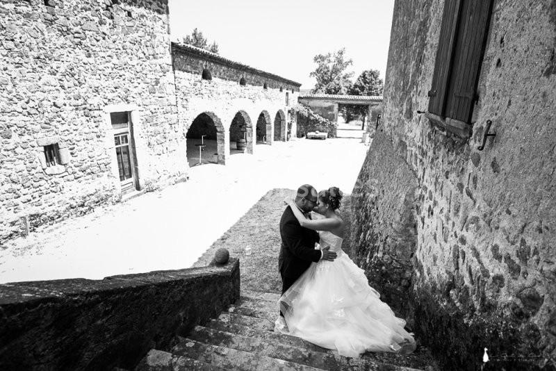 Un couple en tenue de mariage s'embrasse sur des marches en pierre dans une cour ensoleillée bordée de bâtiments anciens.