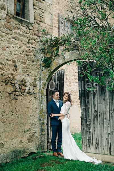 Un couple en tenue de mariage pose devant une porte en bois dans un cadre rustique avec des murs en pierre.