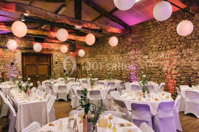 Salle en pierre avec tables rondes dressées pour un événement, décorée de lanternes suspendues et de fleurs.