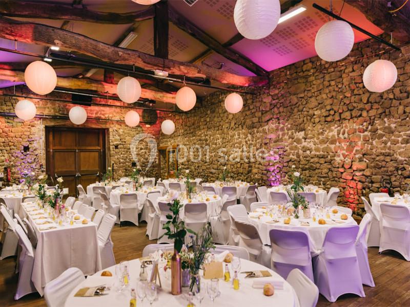 Salle de réception décorée avec des tables rondes dressées, des lanternes suspendues et des murs en pierre apparente.