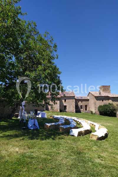 Un jardin verdoyant avec des tables dressées sous un grand arbre, près de bâtiments en pierre sous un ciel bleu.